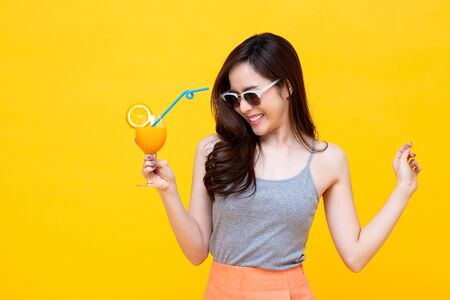Happy Asian woman in summer casual clothes with a glass of fresh fruit juice drink studio shot isolated on colorful yellow backgroundの写真素材