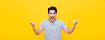 Excited young Asian man raising his fists with smiling delighted face, yes gesture, celebrating success on yellow banner backgroundの写真素材