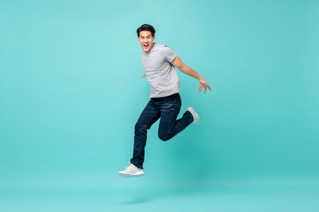 Energetic happy young Asian man in casual clothes jumping, studio shot isolated in light blue backgroundの写真素材