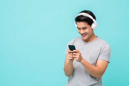 Happy young Asian man wearing wireless headphones listening to music from smartphone studio shot isolated on light blue backgroundの写真素材