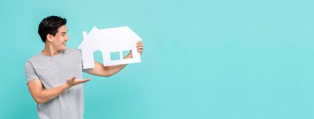 Portrait of young handsome Asian man holding paper home on light blue banner background for real estate conceptの写真素材