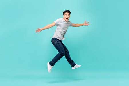 Energetic happy young Asian man in casual clothes jumping, studio shot isolated in light blue...