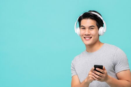 Happy young Asian man wearing wireless headphones listening to music from smartphone studio shot isolated on light blue backgroundの写真素材