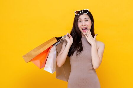 Young Asian woman carrying colorful shopping bags in surprised and excited gesture studio shot isolated on yellow backgroundの写真素材
