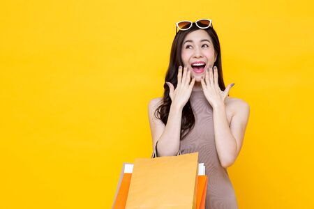 Young Asian woman carrying colorful shopping bags in surprised and excited gesture studio shot isolated on yellow background with copy spaceの写真素材