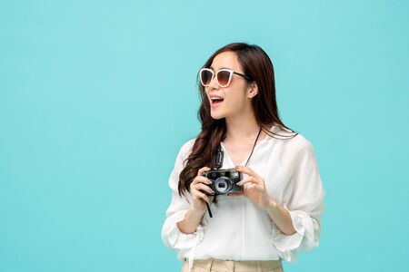 Young amazed female asian tourist in casual white attire holding a camera in sky blue backgroundの写真素材
