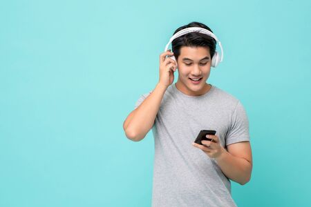Happy young Asian man wearing wireless headphones listening to music from smartphone studio shot isolated on light blue backgroundの写真素材