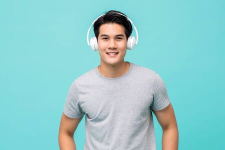 Young smiling handsome Asian man wearing wireless headphones listening to music in light blue backgroundの写真素材