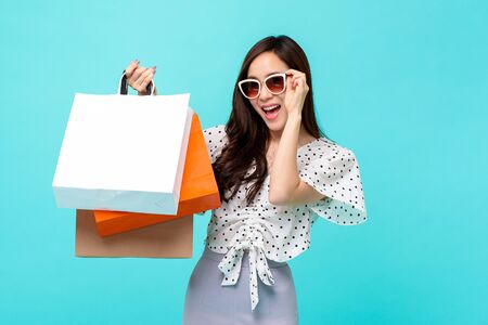Happy beautiful Asian shopaholic woman carrying shopping bags in colorful light blue background, summer sale conceptの写真素材