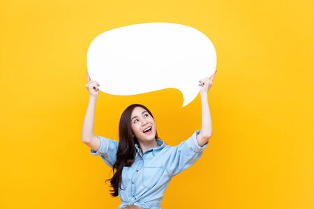 Excited young beautiful Asian woman holding and looking up to speech bubble with empty space for text on colorful yellow backgroundの写真素材