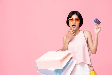 Young pretty Asian woman with shopping bags showing credit card in hand studio shot isolated on colorful pink backgroundの写真素材
