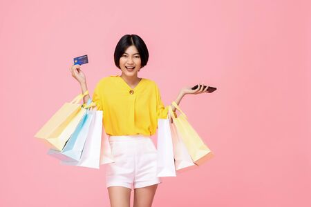Pretty smiling Asian woman with shopping bags showing credit card in hand studio shot isolated on pink backgroundの写真素材
