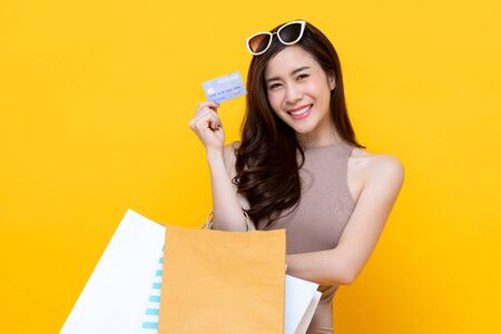 Beautiful smiling Asian woman with shopping bags showing credit card in hand studio shot isolated on colorful yellow backgroundの写真素材