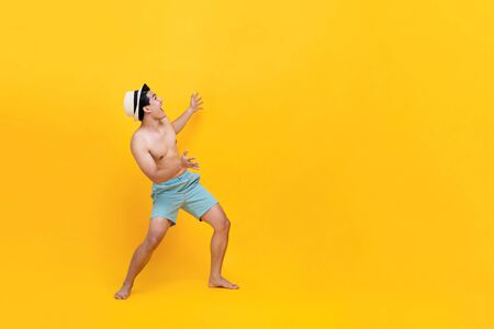 Portrait of enthusiastic shirtless young asian man ready for summer with empty space beside him in a yellow isolated studio backgroundの写真素材