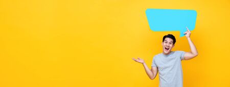 Excited young handsome Asian man holding speech bubble with empty space for text on yellow banner backgroundの写真素材
