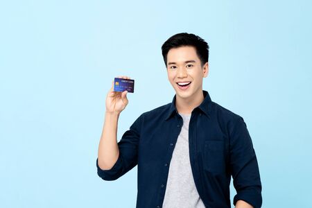 Happy handsome young Asian man showing credit card isolated on light blue backgroundの写真素材