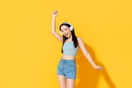 Happy young Asian woman in summer wear listening to music from headphones and dancing isolated on yellow backgroundの写真素材