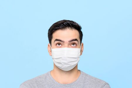 Young man wearing medical face mask thinking and looking up to empty space above isolated on light blue studio backgroundの写真素材
