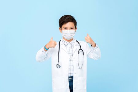 Cute boy doctor wearing medical mask and giving thumbs up isolated on light blue backgroundの写真素材