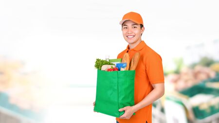 Hygienic smiling Asian delivery man carrying grocery shopping bag in supermarket backgroundの写真素材