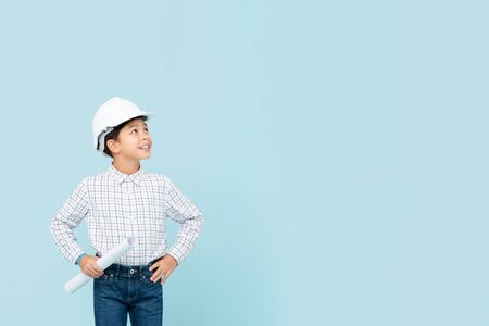 Smiling young asian boy aspiring to be future engineer holding white hard hat and blueprint looking to copy space aside in blue isolated studio backgorundの写真素材