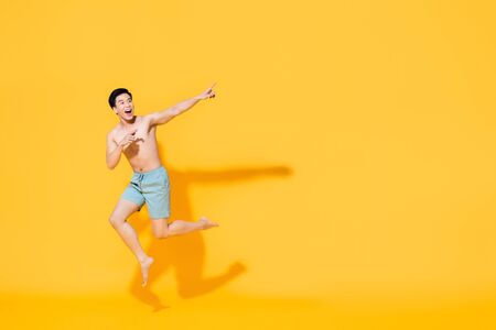 Full length portrait of young happy shirtless Asian man in beach attire jumping in mid-air pointing two fingers at blank space beside in yellow isolated studio backgroundの写真素材