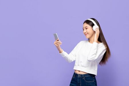 Portrait of young smiling Asian woman listening to music on mobile phone using wireless headphone in purple isolated studio backgroundの写真素材