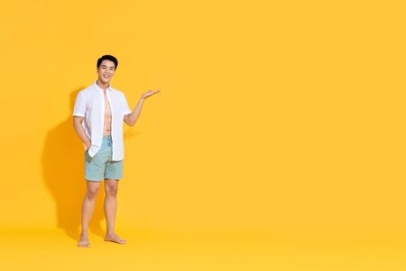 Young smiling handsome Asian man in beach attire doing open palm gesture showing empty space  in summer yellow isolated studio backgroundの写真素材