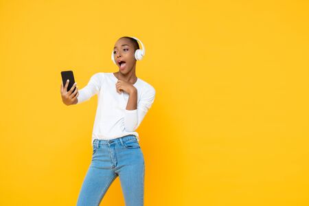 Confident African American woman wearing headphones listening to music from mobile phone in yellow isolated studio backgroundの写真素材