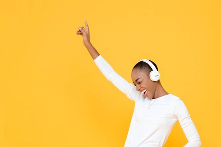 Happy young African American woman raising hand up while listening to music in wireless headphones on yellow isolated studio backgroundの写真素材