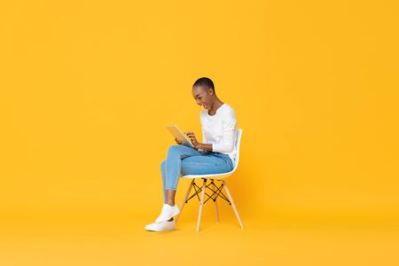 Happy young African American woman sitting on a chair using tablet computer in yellow isolated studio backgroundの写真素材