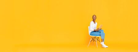 Trendy smiling African American woman sitting on a chair using tablet computer thinking and looking at empty space aside isolated yellow banner backgroundの写真素材