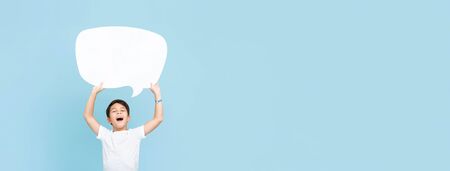 Waist up portrait of smiling Asian boy holding up an empty white speech bubble in light blue banner background with copy spaceの写真素材
