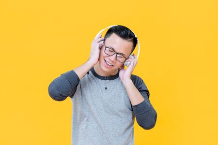 Happy Asian man wearing headphones listening to relaxing music with eyes closed isolated on yellow backgroundの写真素材