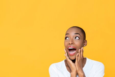 Shocked African American woman with hands on cheeks gasping and looking up to copy space aside isolated on yellow backgroundの写真素材