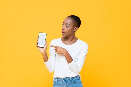 Young African American woman looking and pointing at mobile phone isolated on yellow backgroundの写真素材