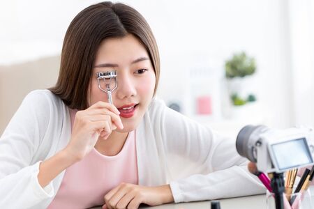 Young Asian girl beauty vlogger testing new eyelash curler and recording video with camera at homeの写真素材