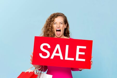 Shocked Caucasian woman carrying shopping bags showing red sale sign isolated on light blue bluebackgroundの写真素材