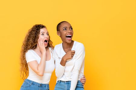 Excited shocked interracial woman friends looking aside isolated on yellow studio backgroundの写真素材