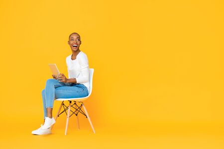 Trendy smiling African American woman sitting on a chair using tablet computer thinking and looking at empty space aside isolated yellow backgroundの写真素材