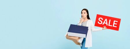 Surprised beautiful Asian woman with shopping bags showing red sale sign isolated on light blue banner background with copy spaceの写真素材