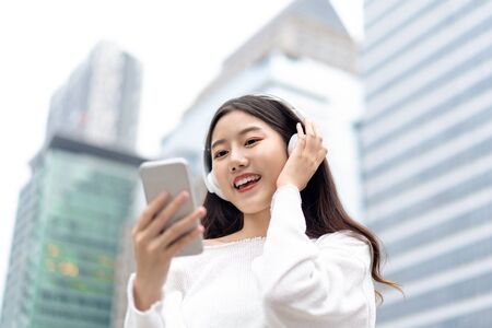 Happy smiling Asian woman wearing headphones and looking at mobile phone while listening to streaming music against city building backgroundの写真素材