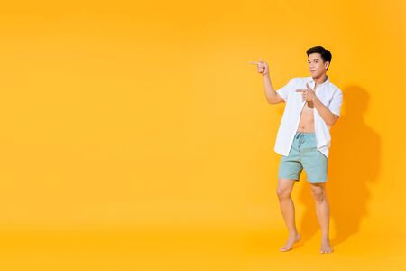 Full body of young handsome Asian man in casual summer outfit pointing hands to copy space aside on colorful yellow backgroundの写真素材