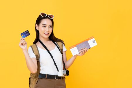 Beautiful Asian tourist woman in yellow background holding passport and boarding pass ready to travel with credit cardの写真素材
