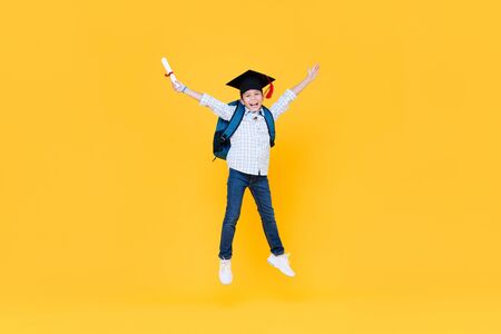 Handsome schoolboy with graduate cap smiling and jumping on yellow background for educatin conceptの写真素材