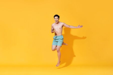 Energetic smiling shirtless handsome Asian man jumping with open hand isolated on yellow backgroundの写真素材