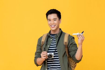 Young handsome smiling Asian tourist man holding plane model and camera isolated on yellow backgroundの写真素材