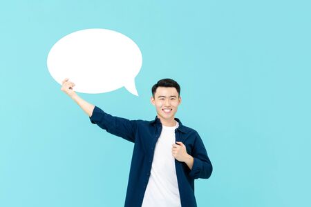 Waist up portrait of young smiling Asian man holding up an empty white speech bubble in light blue studio backgroundの写真素材