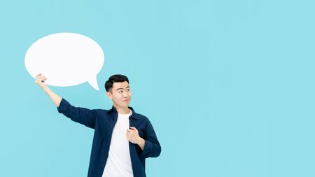 Young smiling Asian man holding empty white speech bubble whlie thinking and looking at copy space in light blue studio backgroundの写真素材