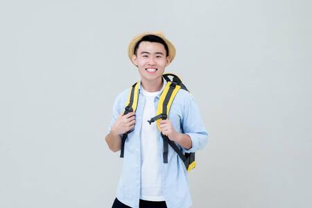 Waist up portrait of young smiling Asian tourist man backpacker holding his yellow backpack straps in gray studio backgroundの写真素材